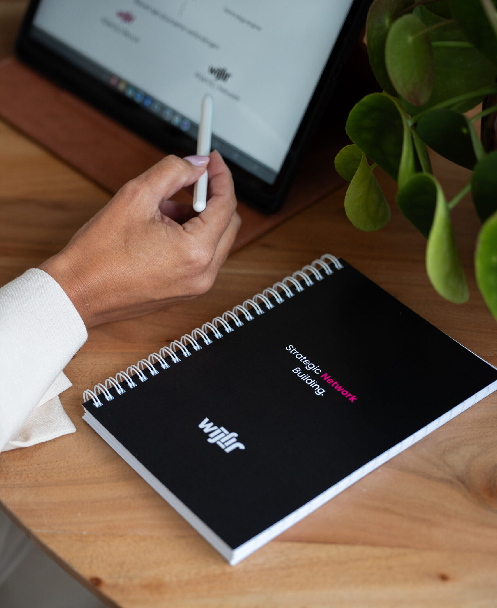 een houten tafel met daarop een tablet, een hand met een tablet pen een een gepersonaliseerde notitieboek van wijenjij